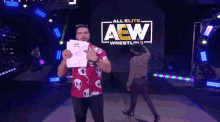 a man in a red shirt is holding a piece of paper in front of a sign that says all elite aew wrestling