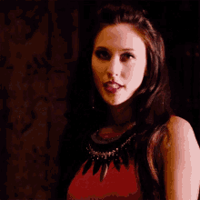 a woman wearing a red top and a black necklace is looking at the camera