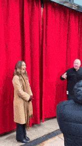a woman in a tan coat is standing in front of a red curtain while a man stands behind her