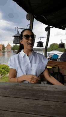 a woman wearing sunglasses laughs while sitting at a table with a lake in the background