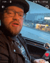a man wearing glasses and a hat is sitting in a car with the time of 7:39 on his phone screen