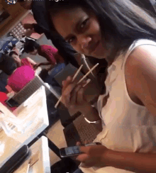 a woman in a white tank top is eating with chopsticks and looking at her phone