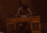 a shirtless man sits at a wooden desk with a piece of paper on it