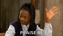 a girl in a school uniform is sitting at a table with her hands in the air and the words `` praise be '' .