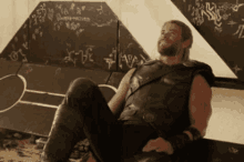 a man with a beard is laying on the ground in front of a wall with graffiti on it that says the