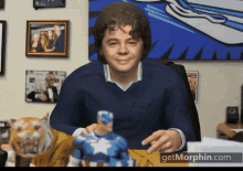 a man in a blue sweater is sitting at a desk with a statue of captain america