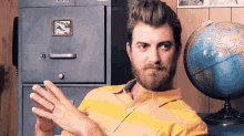 a man with a beard is sitting in front of a filing cabinet and a globe ..
