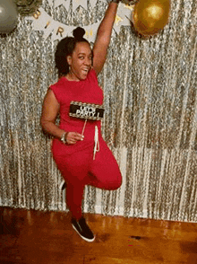a woman in a red jumpsuit is standing on one leg in front of a silver curtain and holding a sign .