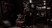 a man in a helmet holds a gun while a baby yoda sits on a box