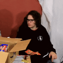 a man wearing glasses is sitting in front of a cardboard box filled with snacks