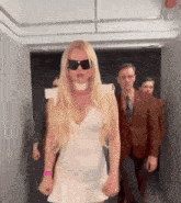 a woman in a white dress and sunglasses is walking down a hallway with men in suits .