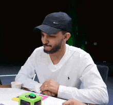 a man wearing a hat and a white shirt is sitting at a table with a green button .