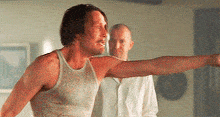 a man in a white tank top is pointing at something while another man stands behind him .
