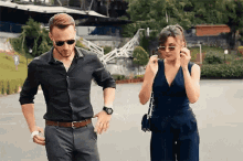 a man and a woman are walking down a street . the woman is wearing sunglasses and the man is wearing a watch .
