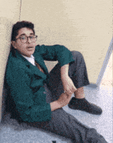 a boy in a green jacket sits on the floor leaning against the wall
