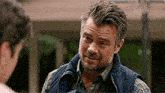 a man with a beard is wearing a vest and talking to another man .