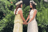 two women wearing flower crowns hold hands in a garden