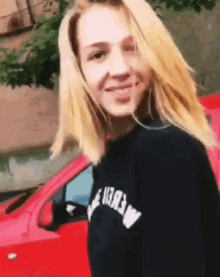 a woman wearing a black sweater with the word merlin on it is standing in front of a red car .