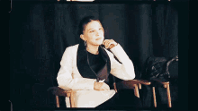 a woman in a white jacket and black vest is sitting in a chair with her hand on her chin .