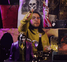 a man in a yellow sweater stands next to a joker figurine and a bottle of wine