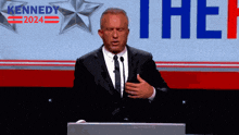 a man in a suit and tie stands at a podium in front of a banner that says kennedy 2024