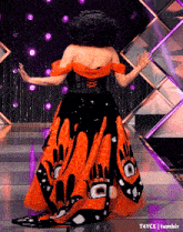 a woman in an orange and black dress is standing on a stage with her arms outstretched .