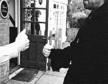a black and white photo of a man giving a thumbs up in front of a door that says front room