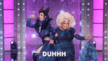 a drag queen is dancing with another drag queen and the word duhhh is on the screen