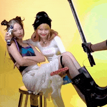 two girls sitting on a stool with one holding a gun and the other holding something