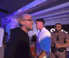 a man wearing glasses is talking to another man in a crowded room