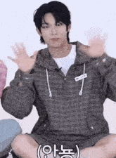 a young man wearing a plaid jacket is sitting on the floor with his hands up .