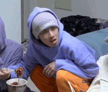 a man wearing a purple hoodie and a beanie with the word lucky on it sits next to a bowl of food