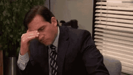a man in a suit and tie is sitting at a desk in front of a window with his hand on his forehead .