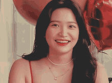 a woman wearing a red top and a necklace is smiling in front of balloons