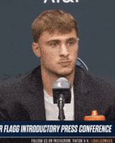 a man in a suit stands in front of a microphone with the words r flagg introductory press conference behind him