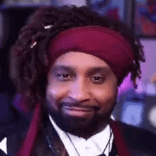a man with dreadlocks and a beard is wearing a red headband and a black jacket .