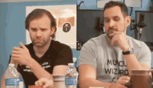 two men are sitting at a table with bottles of water and one has a shirt that says much wizard
