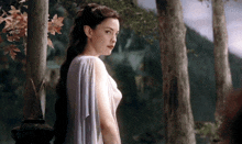 a woman in a white dress is standing in a forest with trees in the background .