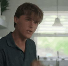 a young man in a blue shirt is standing in a kitchen talking to someone .