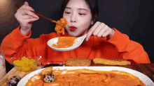 a woman in an orange sweater is eating a plate of food with chopsticks .