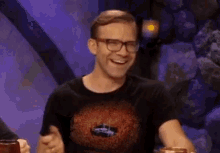 a man wearing glasses is laughing while sitting at a table with a glass of beer .