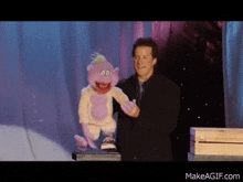 a man is holding a purple puppet on a stage with makeagif.com written on the bottom