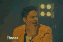 a man in a yellow suit is making a funny face and the word thanos is on the bottom right .