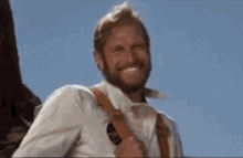 a man with a beard and suspenders is smiling and holding a gun .