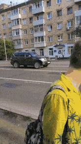 a man in a yellow shirt with palm trees on it looks at a car driving down a street