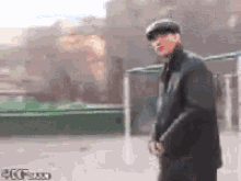 a man wearing a hat and a black jacket is standing in front of a green fence .