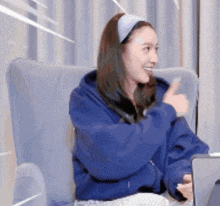 a woman in a blue hoodie is sitting in a chair with a laptop and giving a thumbs up .