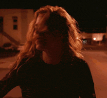 a woman in a black shirt is walking down a street at night with her hair blowing in the wind