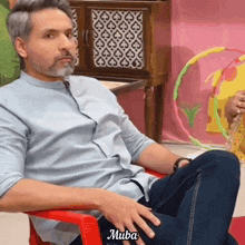 a man in a blue shirt is sitting in a red chair with his hands on his knees and the word muba written on the bottom