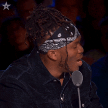 a man wearing a bandana and a denim jacket speaking into a microphone